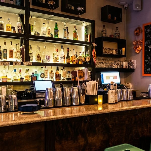 A well-lit bar at Ay Guey! restaurant with liquor bottles, shakers, and a chalkboard menu.