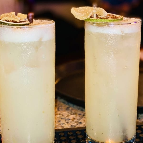 Two tall, frosty cocktails garnished with lime and candied ginger on a speckled bar top.