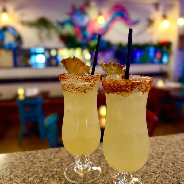 Two pineapple margaritas with chili-lime rims and black straws on a bar, with a colorful mural in the background.
