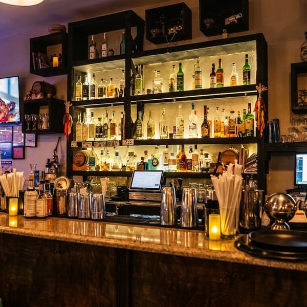 A dimly lit bar with a long wooden counter, liquor bottles on shelves, and bar stools.