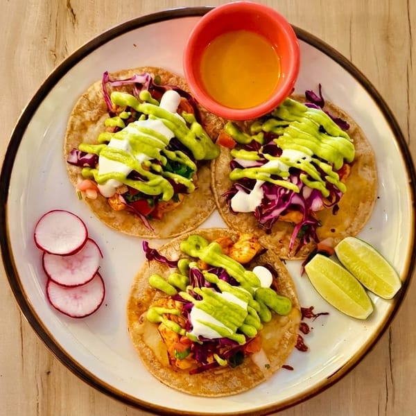 Three vibrant tacos with purple cabbage, white sauce, green drizzle, radishes, and lime wedges on a plate.