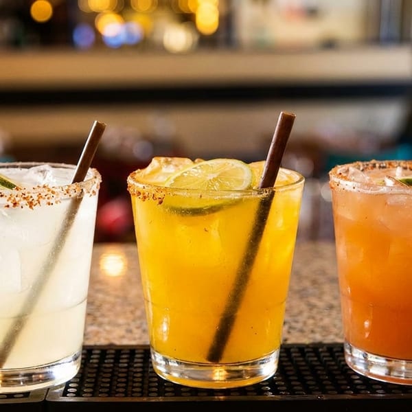 Three colorful margaritas with salt rims, lime garnishes, and straws on a bar.