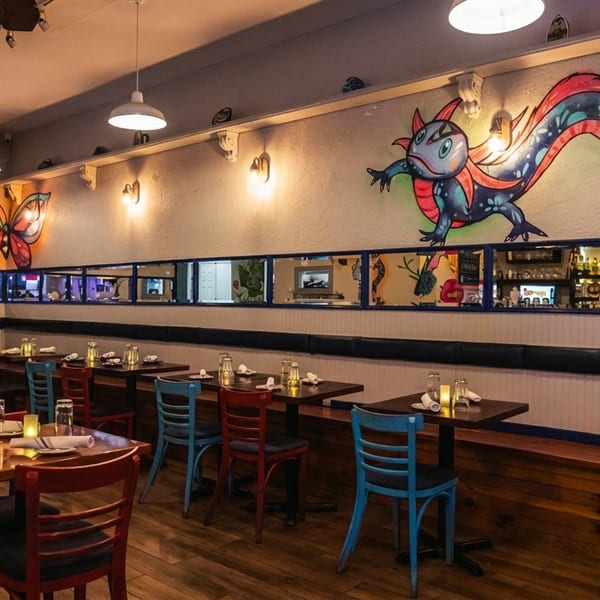 Colorful restaurant interior with murals of a butterfly and lizard, set tables, and a bar reflected in mirrors.