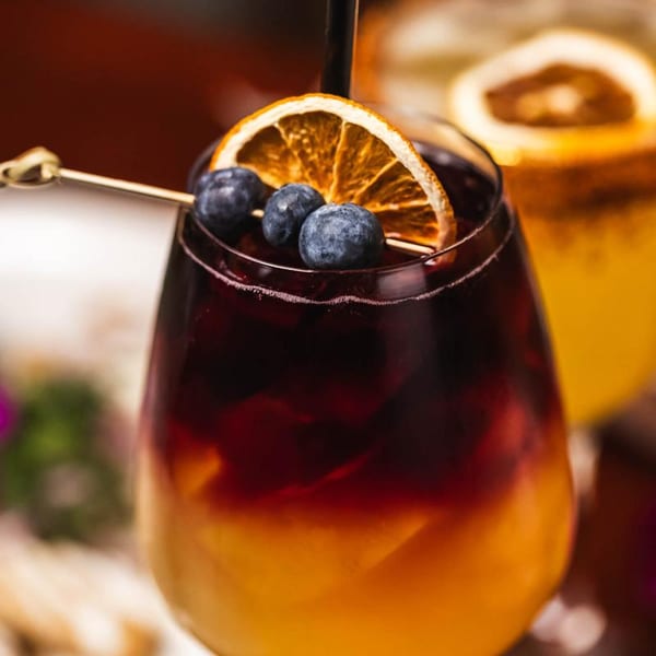 Close-up of a layered cocktail with dark red and orange liquid, garnished with blueberries and a dried orange slice.