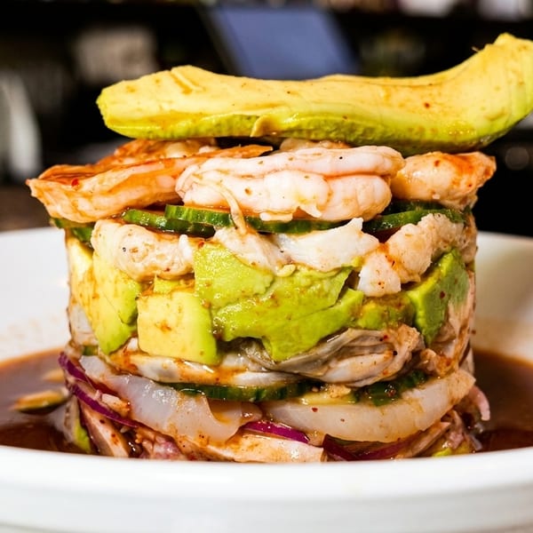 Stacked seafood tower with shrimp, avocado, cucumber, and red onion in a white bowl.