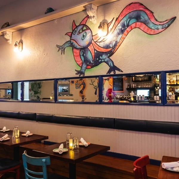 Restaurant interior with wooden tables, colorful chairs, a long banquette, and a vibrant mural above mirrors.