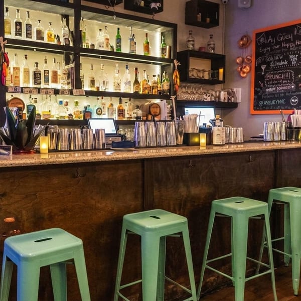 A vibrant bar with a fully stocked liquor shelf, mint green stools, and a colorful hummingbird mural.