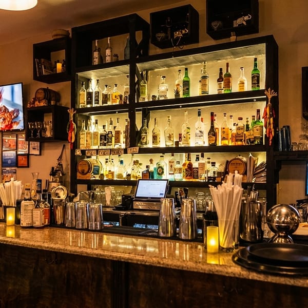 A dimly lit bar with a well-stocked liquor shelf, bar tools, and a TV showing food.